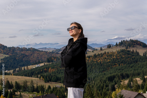 Smiling woman in sunglasses stands in front of a scenic view of the Carpathian Mountains, surrounded by forested hills and autumn colors.