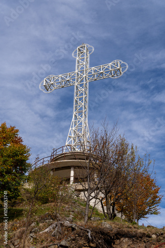 A large metal cross stands on a hilltop surrounded by autumn trees, with a blue sky and wispy clouds in the background.