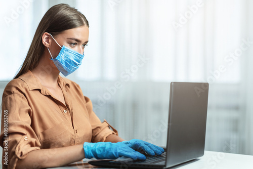 Social Distancing. Side view of girl wearing blue face mask and using laptop at home, sitting at wooden desk photo