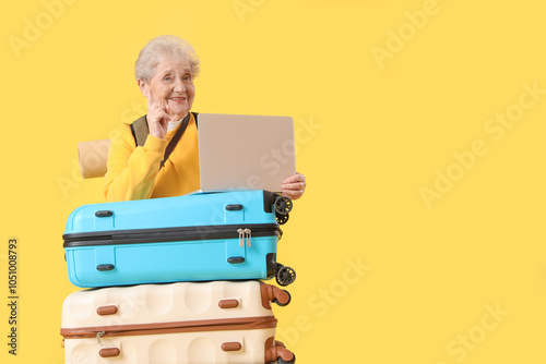 Senior female tourist with laptop and suitcases on yellow background