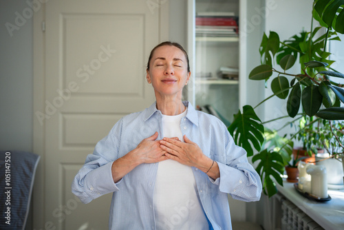 Relaxed smiling woman meditates with hands on chest, practicing breathing exercises at home. Mindfulness practice, focus on breath, self care routine, stress relief, reflection and holistic wellness