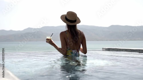 Back view of a woman in swimming costume and sun hat enjoying drink while relaxing on mineral water hot spring in Lesvos Island, Thermi on Yera, Gimbal shot photo