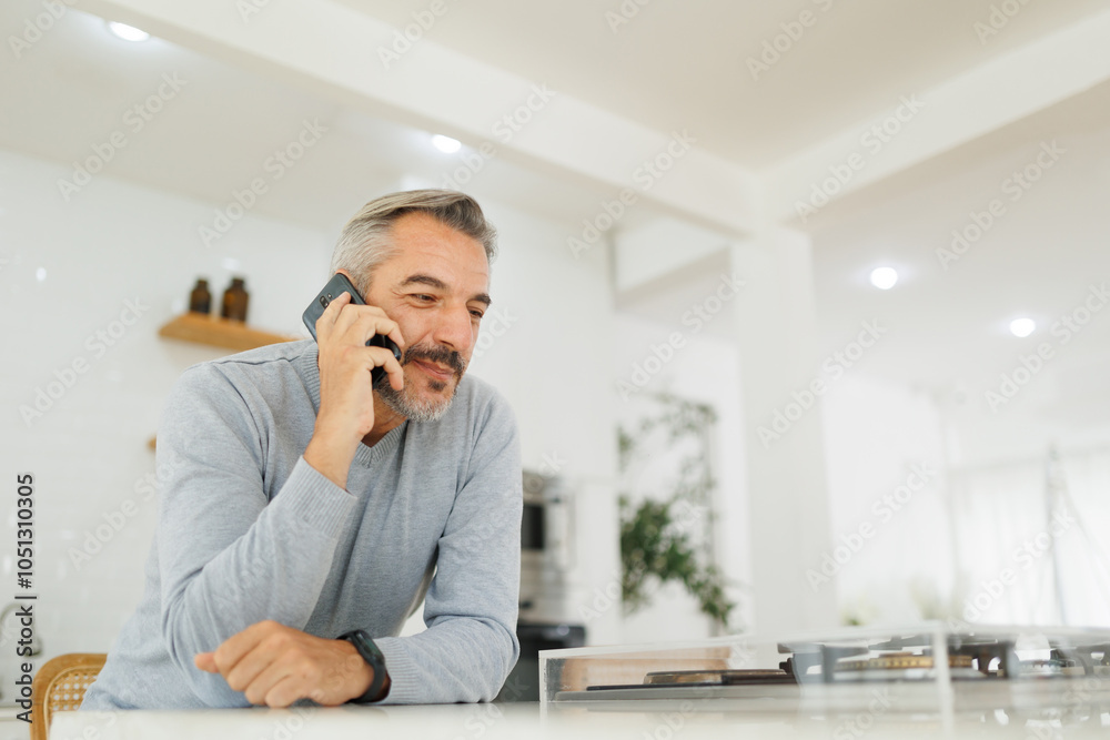 Middle aged man talking on a smartphone with a relaxed expression, leaning on a kitchen counter in a modern kitchen interior. Middle aged man talking on a smartphone with a relaxed expression, leaning on a kitchen counter in a modern kitchen interior.