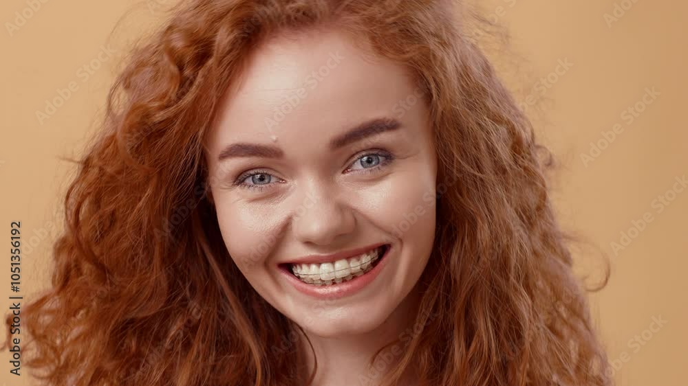 A cheerful young woman with vibrant curly red hair beams joyfully, showcasing her braces. The warm background enhances her lively expression, radiating happiness and positivity. A cheerful young woman with vibrant curly red hair beams joyfully, showcasing her braces. The warm background enhances her lively expression, radiating happiness and positivity.