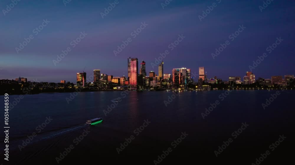 Aerial view of a ferry is traveling through the dark water in front of a city skyline. The city is illuminated by lights, and the sky is a deep blue with a hint of purple. Aerial view of a ferry is traveling through the dark water in front of a city skyline. The city is illuminated by lights, and the sky is a deep blue with a hint of purple.