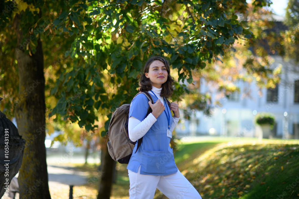 Fototapeta premium Beautiful nurse walking to work with backpack on her back. Autumn walk from hospital to home for young healthcare worker.