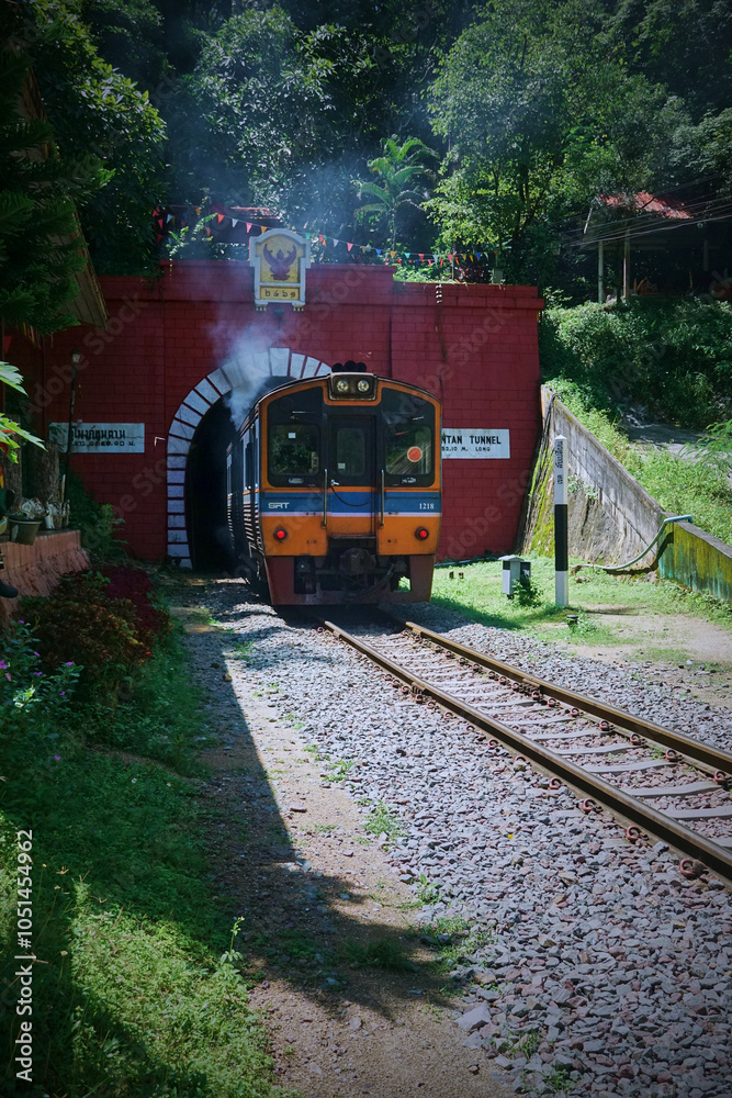 Obraz premium A blue and orange train entering a red brick tunnel surrounded by lush greenery.