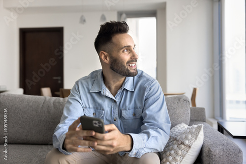 Dreamy positive handsome Latin man using gadget for online communication at home, holding smartphone, looking at window away, smiling, thinking on Internet technology on gadget for work