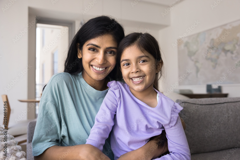 Happy young Indian mother holding sweet daughter kid on lap, sitting on home couch, looking at camera, smiling, hugging child with love, affection, tenderness, promoting motherhood Happy young Indian mother holding sweet daughter kid on lap, sitting on home couch, looking at camera, smiling, hugging child with love, affection, tenderness, promoting motherhood