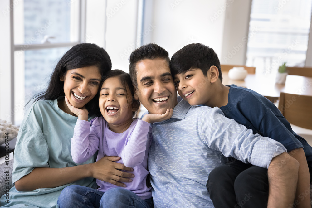 Happy excited Indian parents and two kids having fun at home, sitting close on couch, hugging, laughing, holding younger child in arms, enjoying homey family leisure together Happy excited Indian parents and two kids having fun at home, sitting close on couch, hugging, laughing, holding younger child in arms, enjoying homey family leisure together