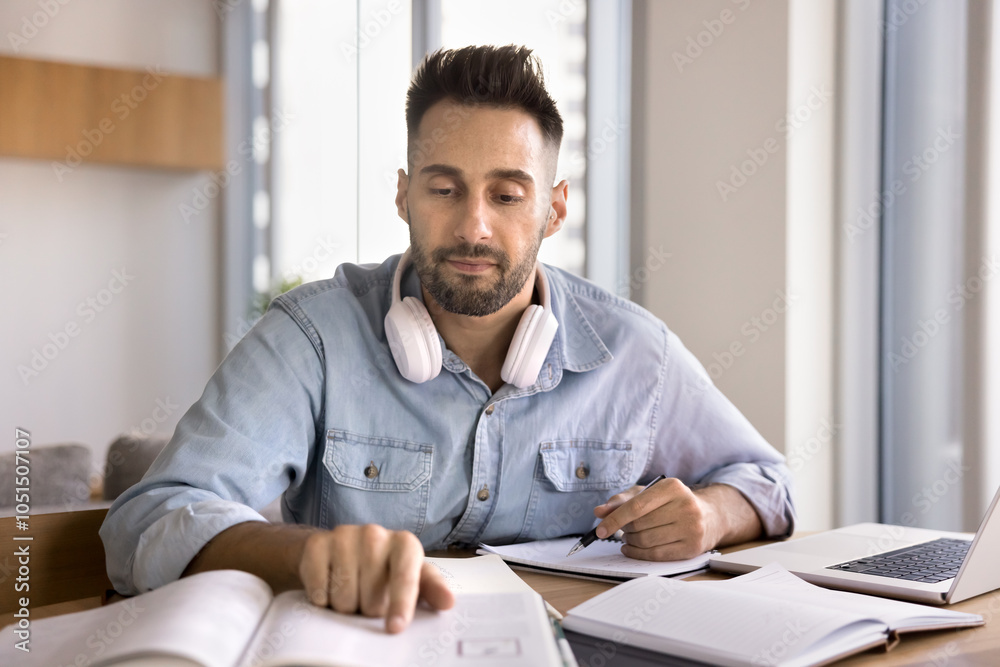 Fototapeta premium Focused adult Latin student man reading learning textbook at home workplace table with laptop, studying for new occupation, preparing for university test, exam, pointing finger at page