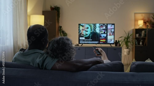 Rear dolly out shot of loving diverse spouses sitting on couch and watching TV after work in evening, husband hugging wife from behind while she choosing movie on streaming service