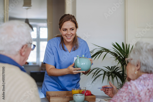 Happy healthcare worker holding teapot and talking to senior couple at home photo