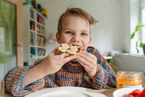 Boy eating bread with sliced bananas at table in home