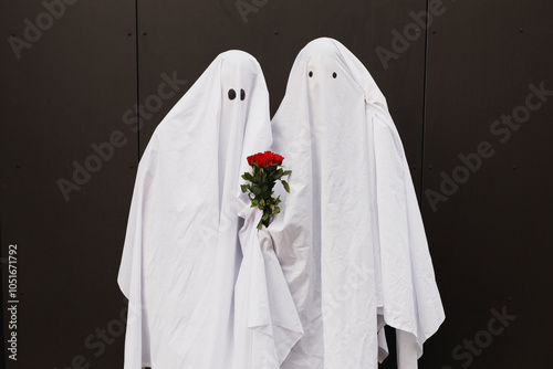Young couple wearing ghost costumes and standing with flowers in front of wall
