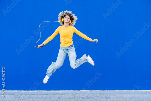 Young woman with headphones jumping in front of blue wall
