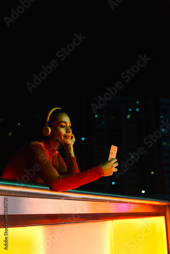 Woman with headphones and hand on chin taking selfie through smart phone at night