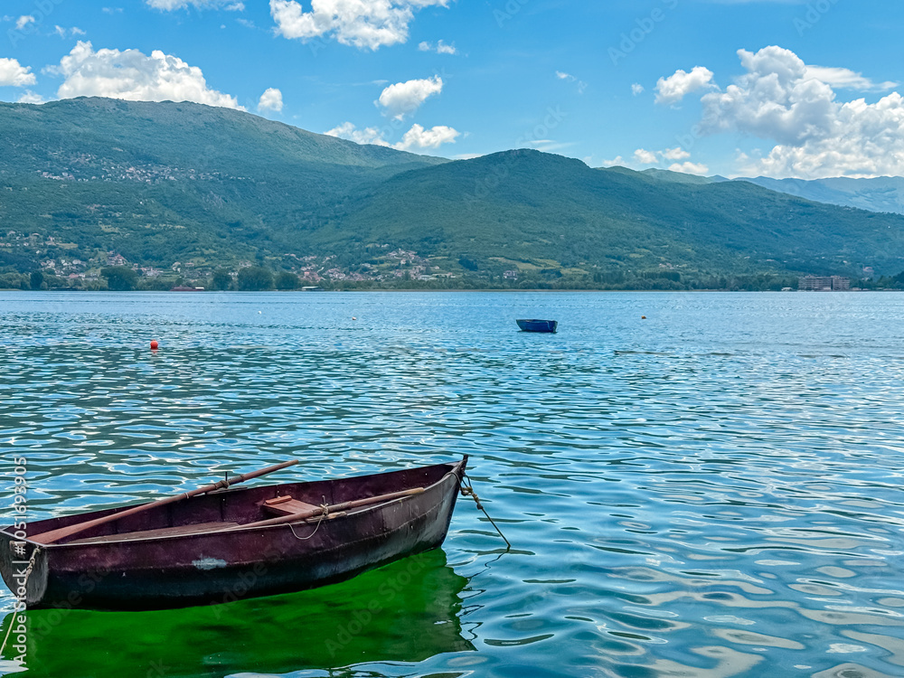 Idyllic view of small boat floating on calm Lake Ohrid in North Macedonia. Rolling hills covered in lush green vegetation. Tranquil peaceful lakeside scene. Summer travel destination in the Balkans Idyllic view of small boat floating on calm Lake Ohrid in North Macedonia. Rolling hills covered in lush green vegetation. Tranquil peaceful lakeside scene. Summer travel destination in the Balkans
