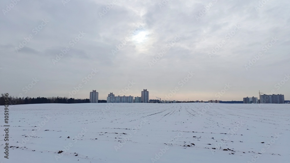 After a snowfall, snow lies on the ground of an agricultural field with trees and buildings behind it. There is a crane and a boiler room pipe behind the buildings. The sun breaks through the clouds After a snowfall, snow lies on the ground of an agricultural field with trees and buildings behind it. There is a crane and a boiler room pipe behind the buildings. The sun breaks through the clouds