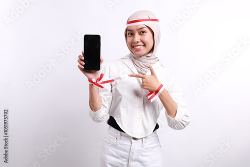 Smiling Happy young Asian Muslim woman pointing a finger at the display mobile phone blank screen, recommends apps isolated over white background. Celebrate Indonesian independence day on 17 August photo