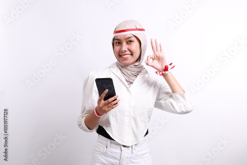 Smiling happy young Asian Muslim woman wearing a hijab holding mobile phone and showing okay sign isolated over white background. Celebrate Indonesian independence day on 17 August photo