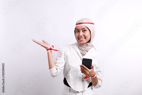 A happy Asian muslim woman wearing red white headband, holding her phone, and pointing copy space beside her, isolated by white background. Indonesia's independence day photo