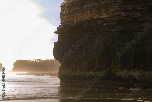 View of a high cliff rising on a beach with black volcanic sand. In the background is the setting sun, with warm rays penetrating the rock. A serene and deserted beach without people. photo