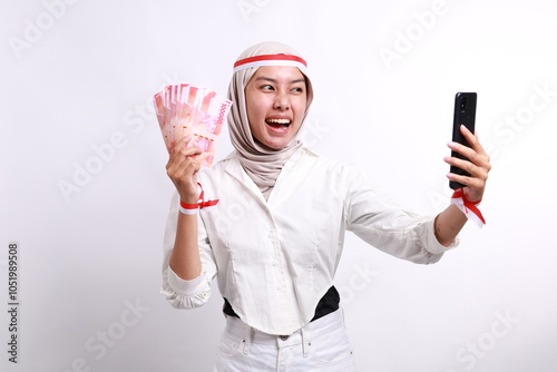 Excited and confident young Asian muslim woman holding mobile phone and indonesia rupiah money banknotes, celebrating Indonesia independence day on August 17 isolated on white background photo