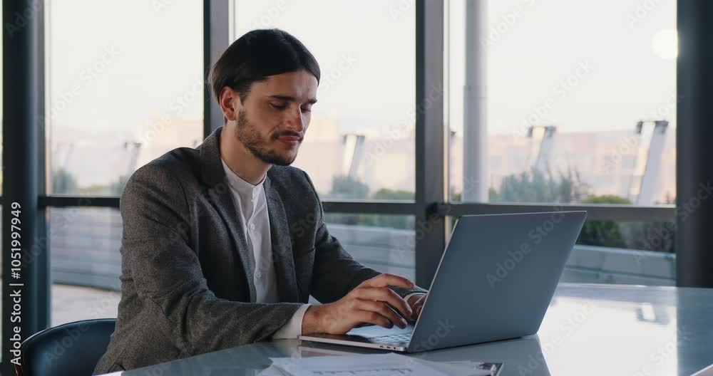 A young professional in a good mood, dressed in a business suit, works on his laptop in a large modern office. Perfect for showcasing productivity, focus, and a positive work environment. A young professional in a good mood, dressed in a business suit, works on his laptop in a large modern office. Perfect for showcasing productivity, focus, and a positive work environment.