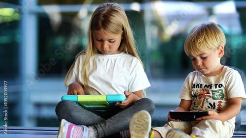 Two young children sitting together using tablets, one smiling at her device, the other focused on his screen.
