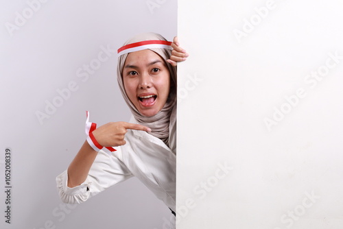 Portrait of young muslim woman with red white flag ribbon happy excited positive smile point finger empty space billboard ad advice choice isolated over white background photo