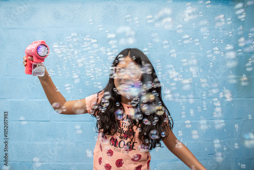 Uma menina, alegre, brincando com uma pistola de fazer bolhas de sabão, com fundo azul. photo