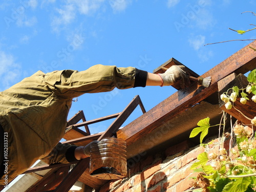a man in work clothes stands on an iron ladder and applies an oily liquid with a brush to a wooden beam at the base of the roof of a brick extension to a house, impregnation of load-bearing roof beams