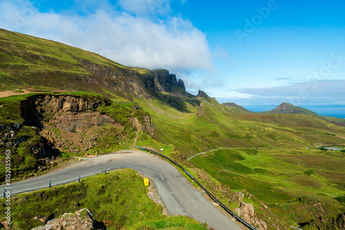 A winding road navigates the dramatic slopes of the Quiraing, Isle of Skye, revealing stunning views of the Scottish Highlands and a distant glimpse of the sea.