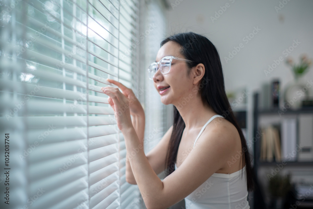 Fototapeta premium Young woman wearing glasses opening blinds and looking out the window