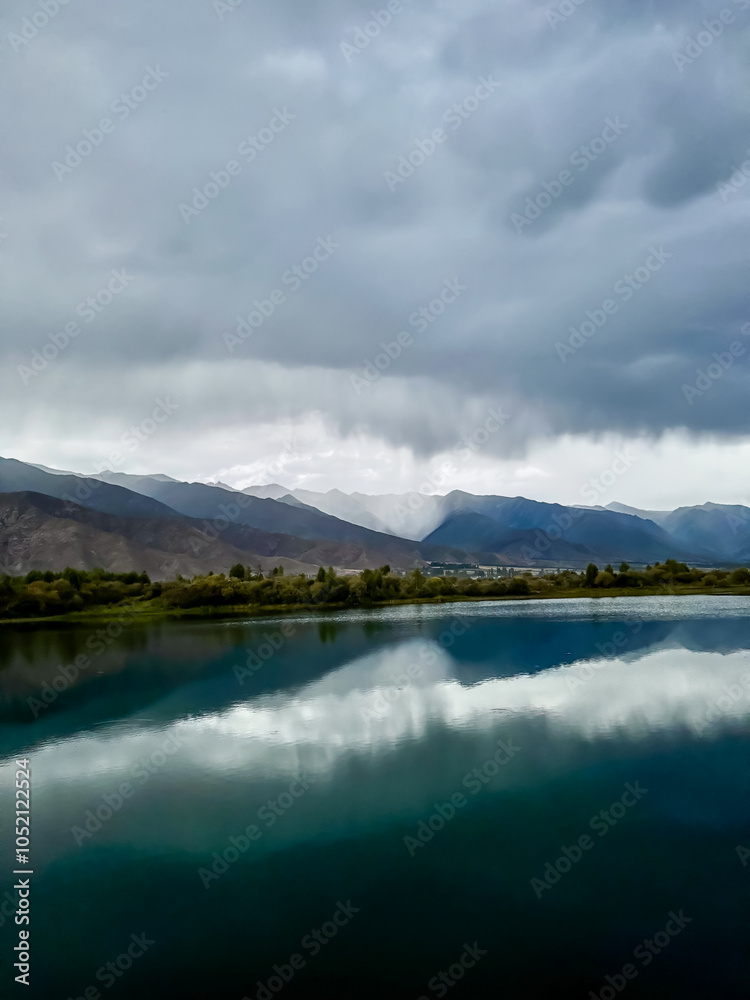 Calm lake waters reflect dark clouds and mountains in the background. The grey sky hangs over the lake, creating a pre-storm atmosphere. Calm lake waters reflect dark clouds and mountains in the background. The grey sky hangs over the lake, creating a pre-storm atmosphere.