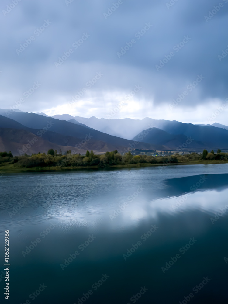 Calm lake waters reflect dark clouds and mountains in the background. The grey sky hangs over the lake, creating a pre-storm atmosphere. Calm lake waters reflect dark clouds and mountains in the background. The grey sky hangs over the lake, creating a pre-storm atmosphere.