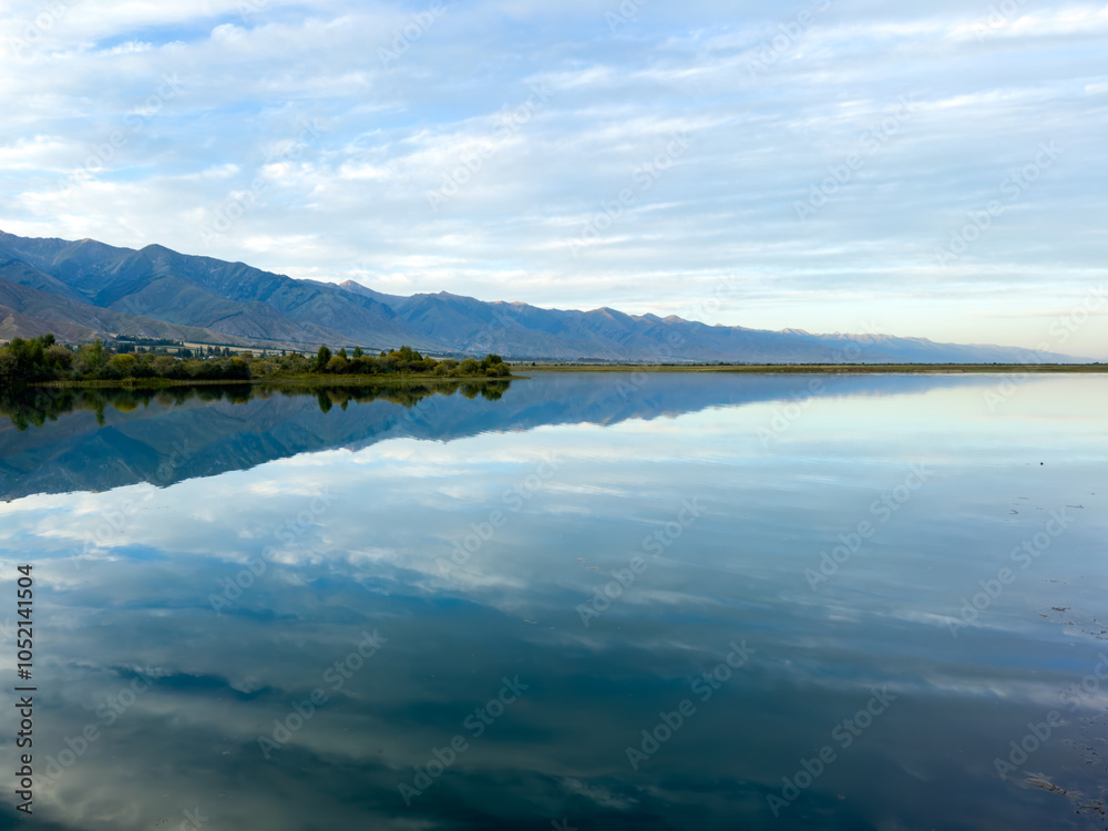 Obraz premium The calm water of a mountain lake reflects the blue sky and white clouds. The green shoreline frames the lake, creating a sense of harmony and tranquility.