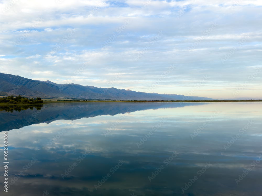 The calm waters of Issyk-Kul lake reflect dramatic clouds and mountains. Silence and tranquility in a natural oasis in Kyrgyzstan The calm waters of Issyk-Kul lake reflect dramatic clouds and mountains. Silence and tranquility in a natural oasis in Kyrgyzstan