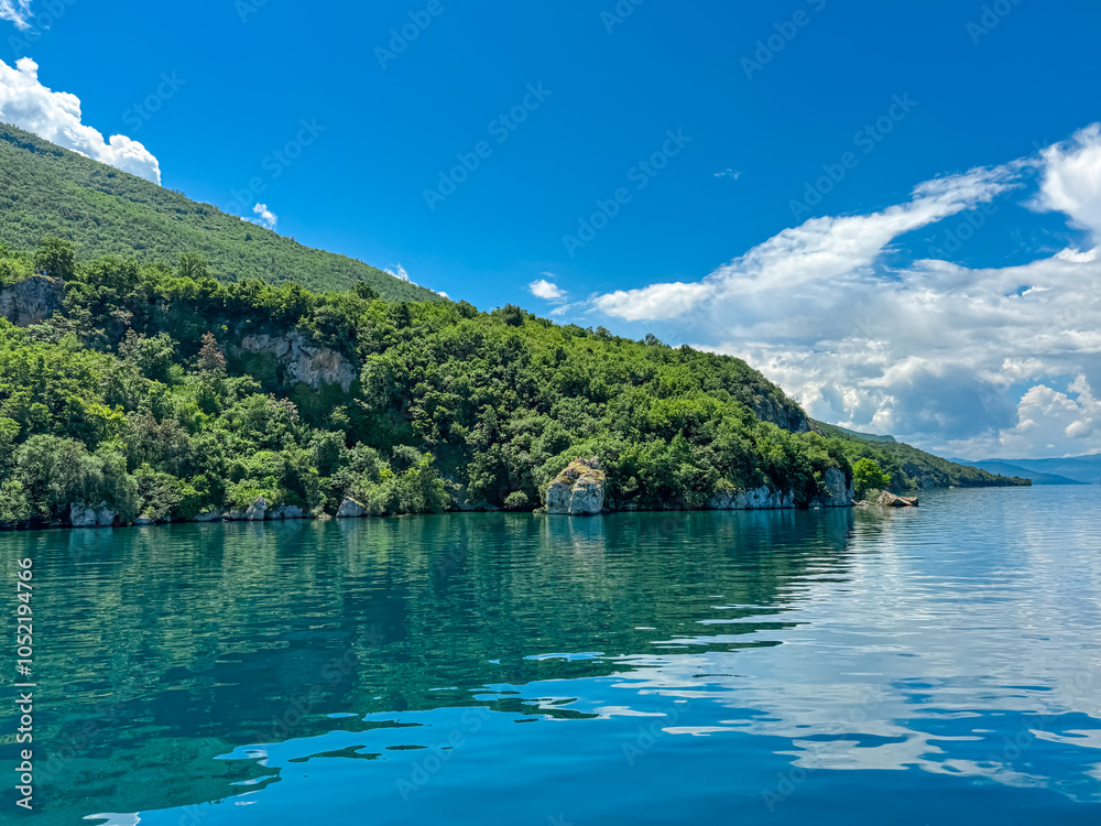 Obraz premium Serene lakeside scene along the shoreline of Lake Ohrid, North Macedonia. Mountain range of National Park Galicica in the distance. Slopes and hills covered with lush green vegetation and forest