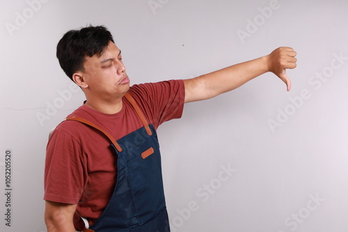 Side view of Discontent Asian man wearing blue apron shows disapproval sign, keeps thumb down, expresses dislike, frowns face in discontent, isolated white background. Body language concept. photo