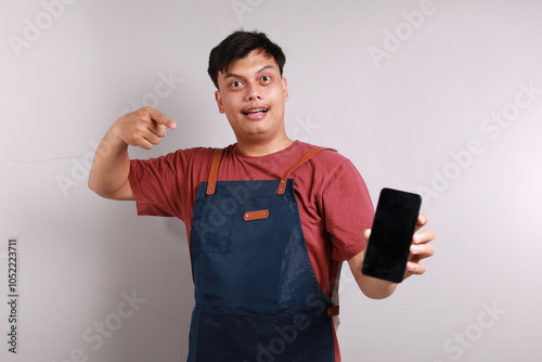 Young handsome asian barista man wearing apron pointing finger at mobile screen, showing app and smiling, standing over white background. Selective focus