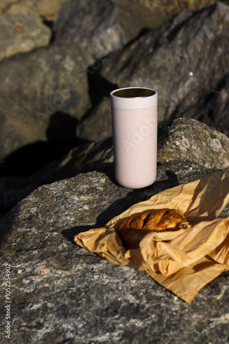 A cozy outdoor scene featuring a thermos mug and a freshly baked pastry resting on rugged Norwegian rocks under clear, sunny skies. The stainless steel mug gleams in the sunlight.