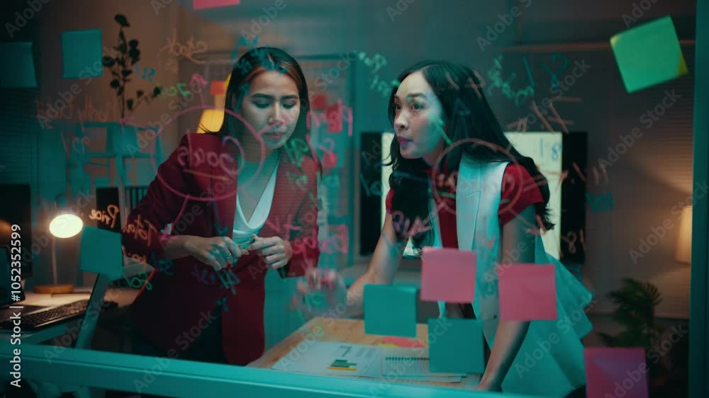 Two young businesswomen are discussing and brainstorming, using a glass wall as a whiteboard. They are writing with a marker and using colorful sticky notes