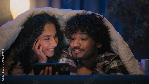 Multiracial couple man and woman happy cover with blanket under duvet at night evening home using mobile phone online browsing talking discuss video call shopping with smartphone family gadget addict