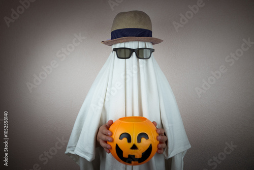 A person in a Halloween ghost costume, a capelus and sun-drenched eyepieces, trims a watermelon against a gloomy dark background. More dramatic photo.
