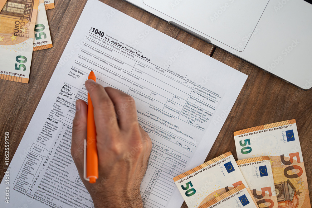 Fototapeta premium Hand filling out a 1040 tax form with an orange pen on a wooden desk surrounded by euro bills and a laptop. Personal finance, tax preparation, and accounting concept 