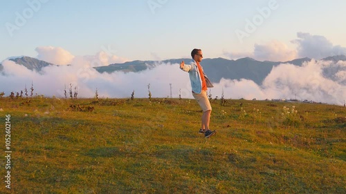 Man stands on grassy mountainside with his arms outstretched, embracing beauty of surrounding clouds and mountains. He dances and enjoys life with smile. Retarded motion photo