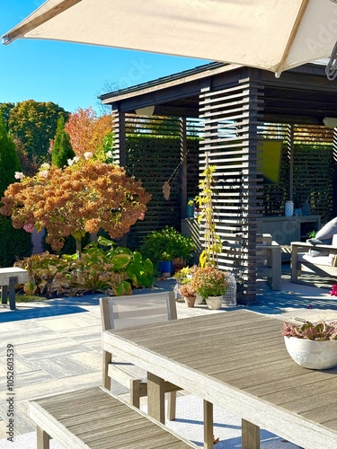 High-end outdoor living space in an unseasonably warm autumn.  A backyard patio with pergola, lounge chair, rug, bar, dining set, couch, television, pool waterfall, umbrella, fire pit and grey patio. photo