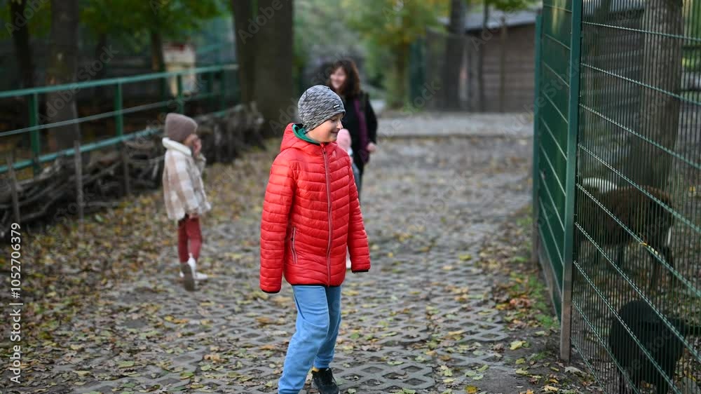A family of a mother and three children visiting a zoo in autumn. The boy is wearing a red jacket and the girls are wearing hats. They are looking at animals behind a fence.